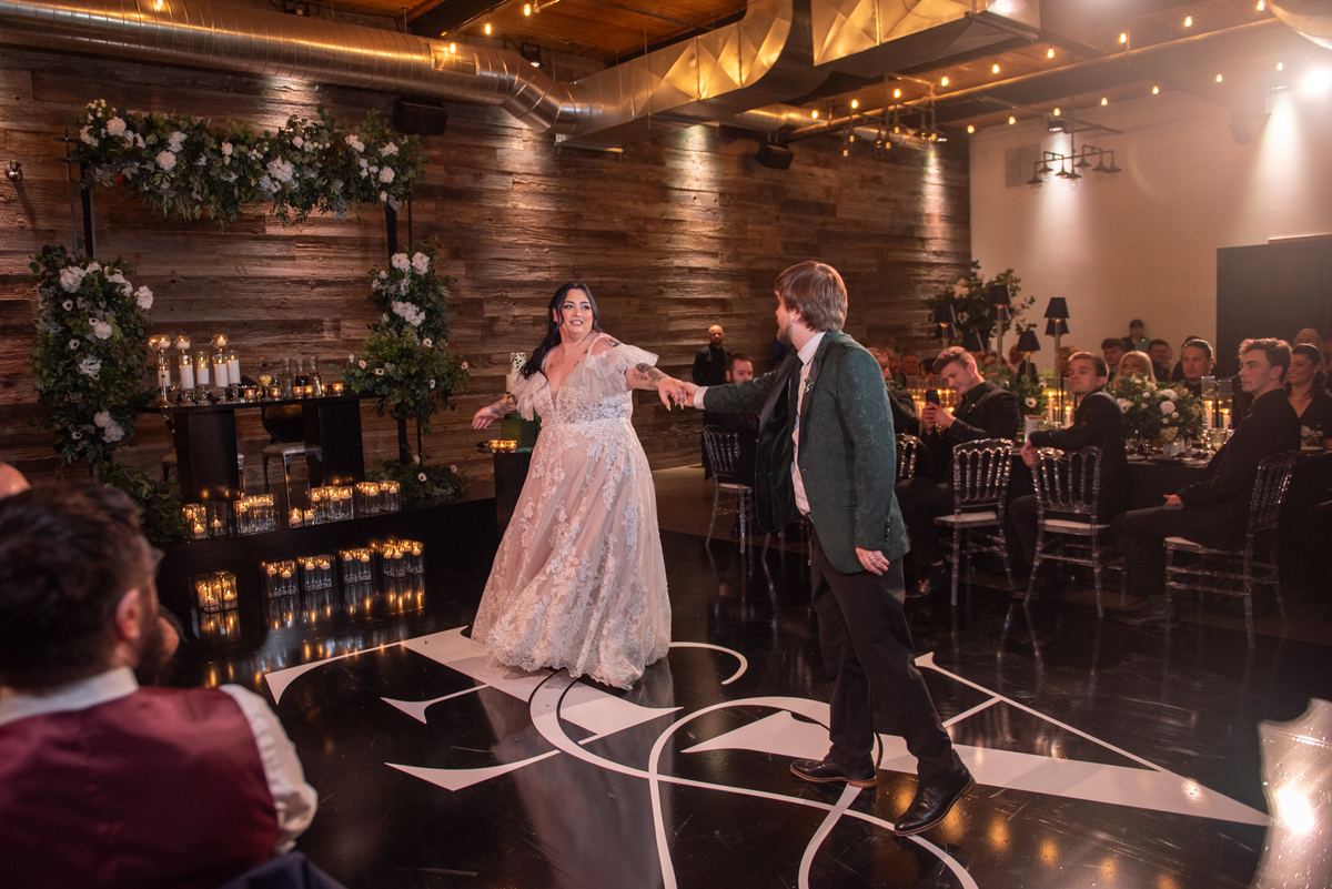 Bride and groom dance joyfully on black-and-white floor, surrounded by candlelit tables and rustic decor.