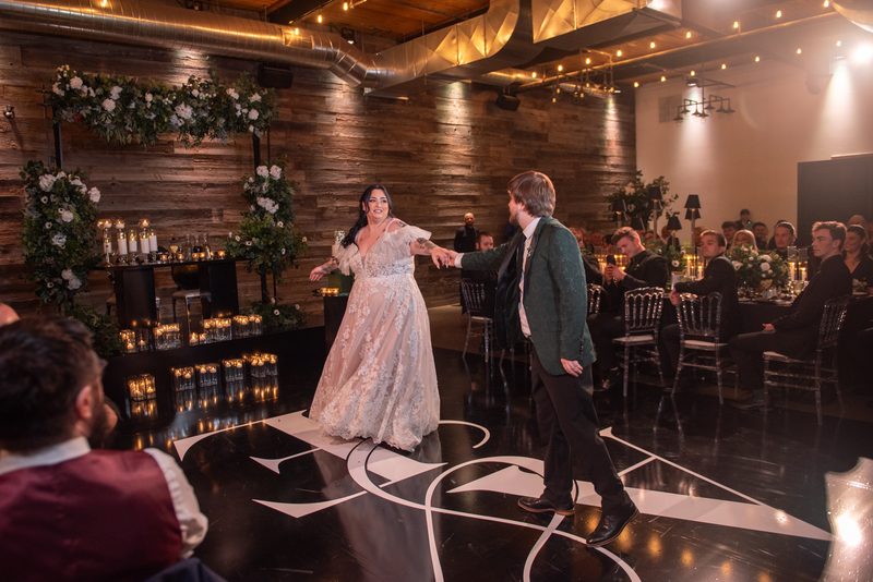 Bride and groom dance joyfully on black-and-white floor, surrounded by candlelit tables and rustic decor.