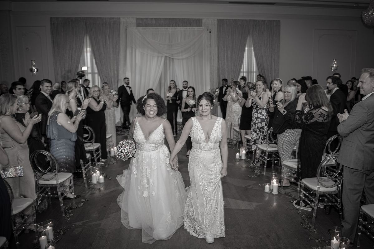 Two brides in elegant gowns walk down the aisle hand-in-hand as guests applaud and candles illuminate the scene.
