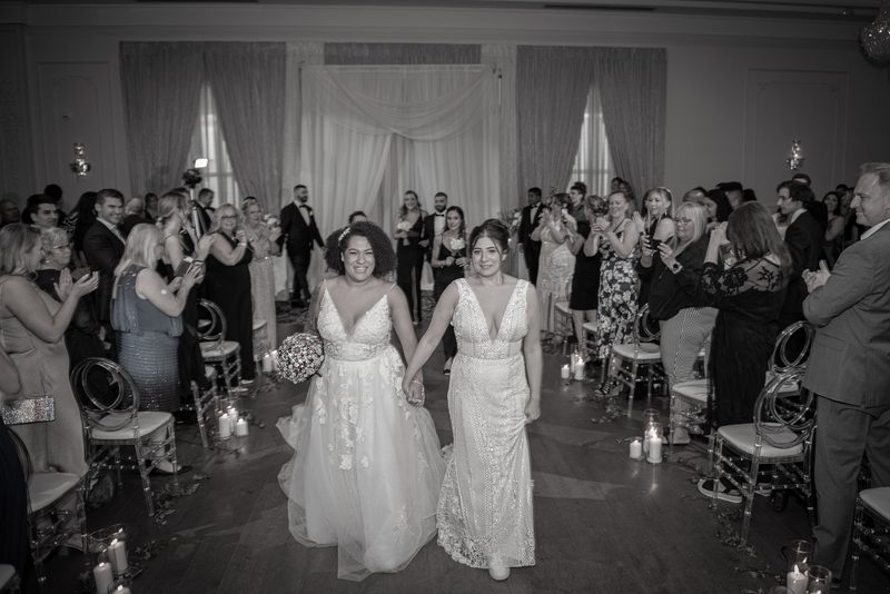 Two brides in elegant gowns walk down the aisle hand-in-hand as guests applaud and candles illuminate the scene.