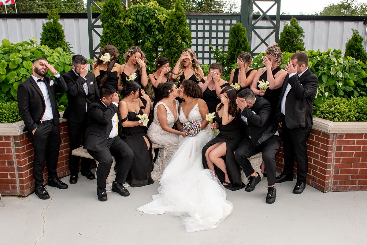 Two brides kiss joyfully with their wedding party reacting playfully, covered in greenery and brick planters.