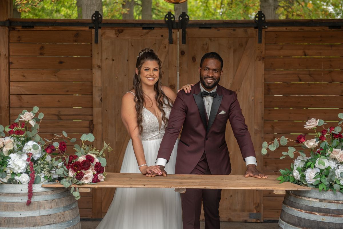 Bride and groom smile at camera near wooden barn door with wine barrel flowers in rustic vineyard setting.
