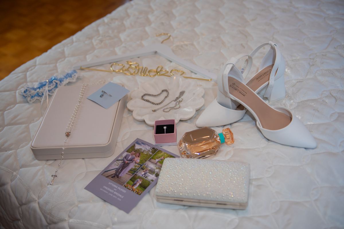 A bride's wedding day items including shoes, clutch, rosary, perfume, and name card lie on a white quilt.