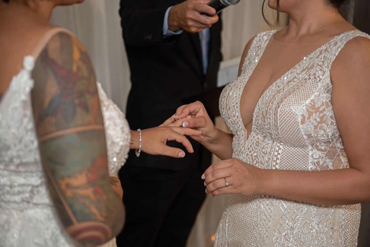 A bride places a ring on her partner's finger during an indoor wedding ceremony, with an officiant nearby.