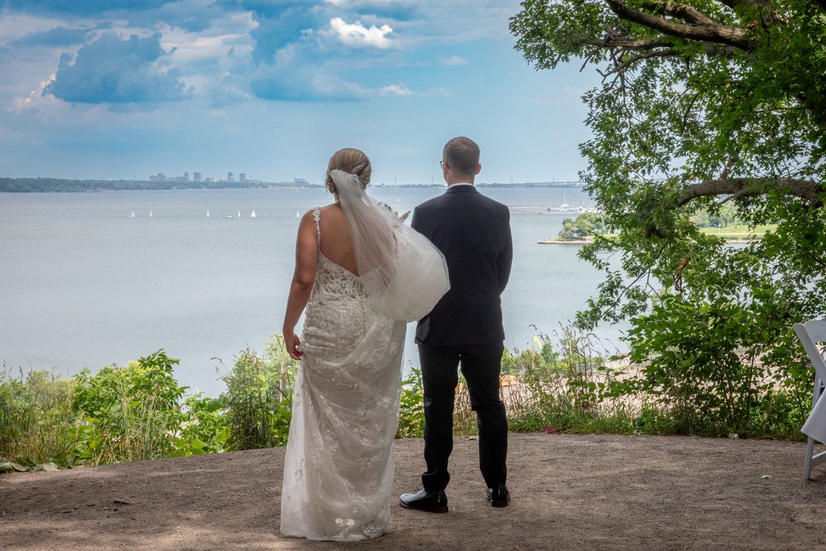 A bride and groom stand on a path by water, gazing at sailboats and city skyline under partly cloudy sky.