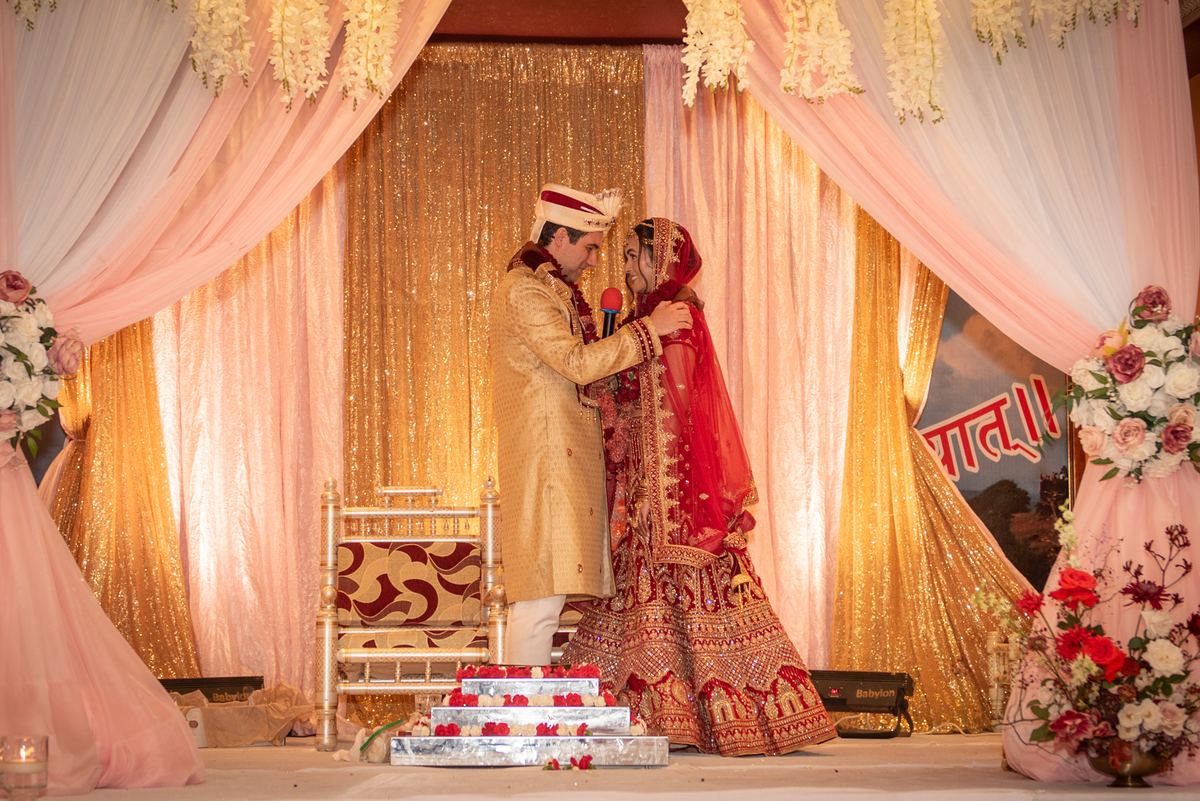 A bride in a red lehenga and a groom in a golden sherwani embrace on stage, holding microphones near floral arrangements.