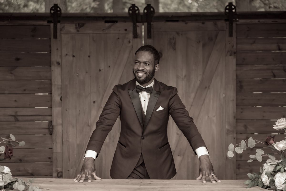 A smiling groom in a tuxedo leans against a wooden table near rustic barn doors, illuminated by soft light.