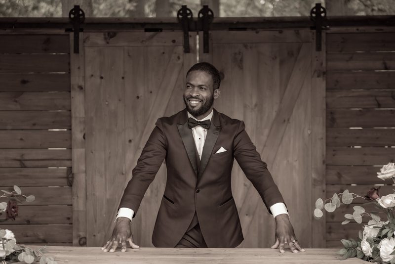 A smiling groom in a tuxedo leans against a wooden table near rustic barn doors, illuminated by soft light.