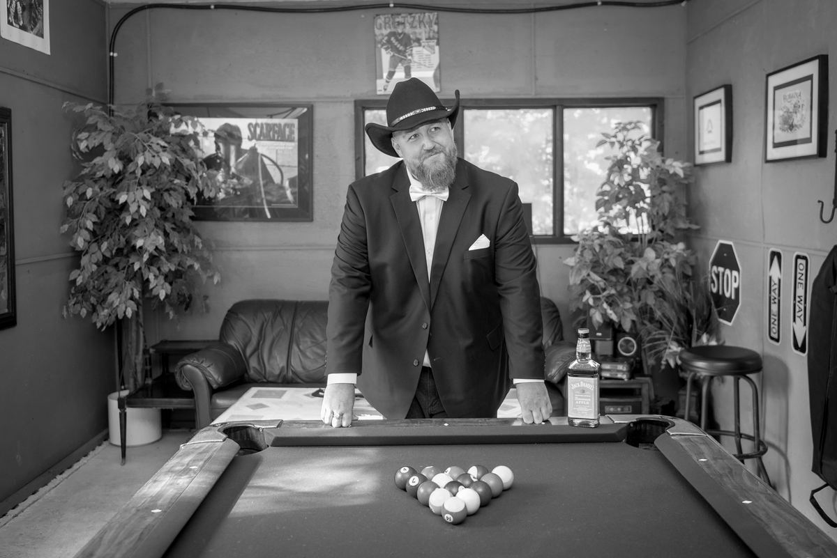 A man in a suit and cowboy hat smirks confidently behind a pool table in a decorated room with movie posters.