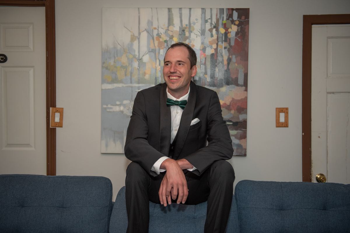 A man in a black tuxedo with a green bow tie sits on a blue couch, smiling next to an autumn tree painting.