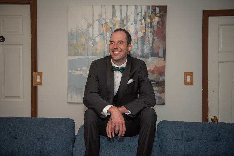 A man in a black tuxedo with a green bow tie sits on a blue couch, smiling next to an autumn tree painting.
