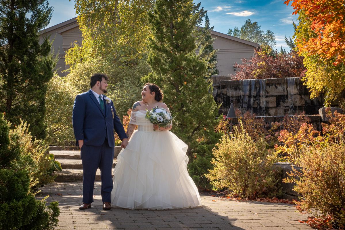 A smiling bride and groom in formal attire stand on a stone path, surrounded by autumn foliage and a stone waterfall.