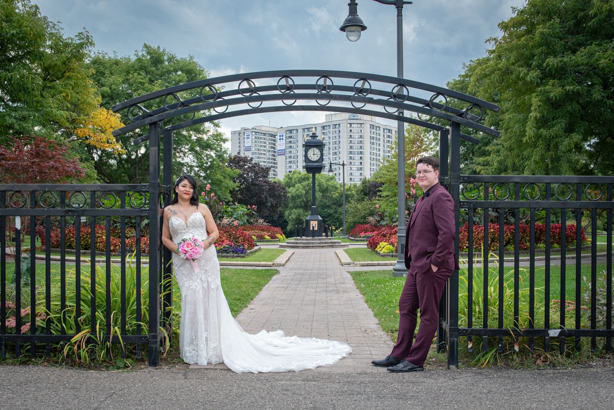 A bride in white and groom in maroon pose at a wrought-iron gate in a park with a clock tower and cityscape visible.