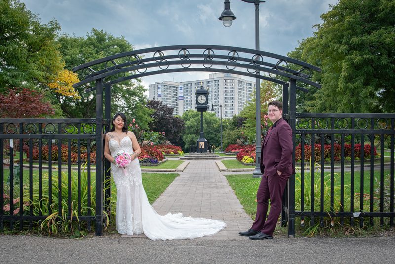 A bride in white and groom in maroon pose at a wrought-iron gate in a park with a clock tower and cityscape visible.