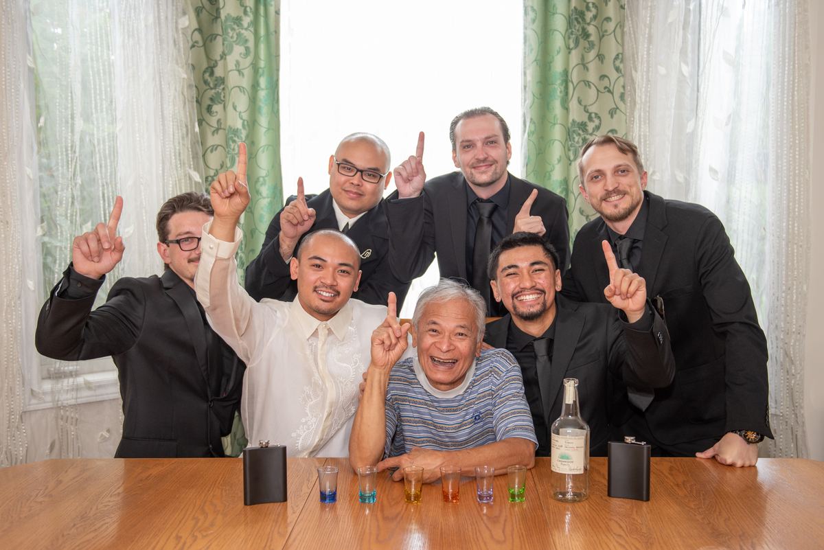 Seven men, including an elderly man at the center, raise a finger in unison around a table set with shot glasses and flasks.