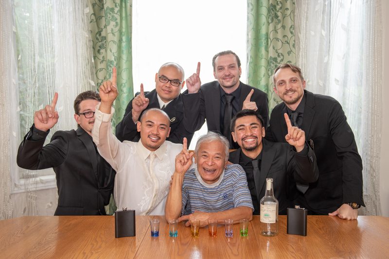 Seven men, including an elderly man at the center, raise a finger in unison around a table set with shot glasses and flasks.