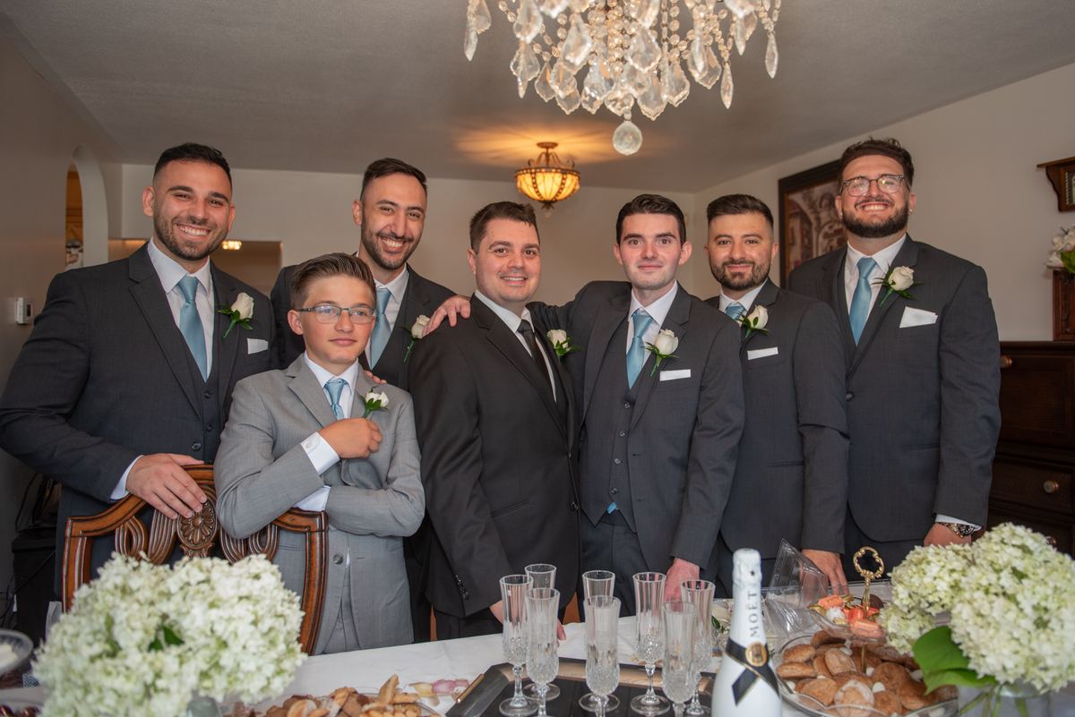 Groomsmen and groom pose in formal suits with boutonnieres under warm lighting during a celebration.