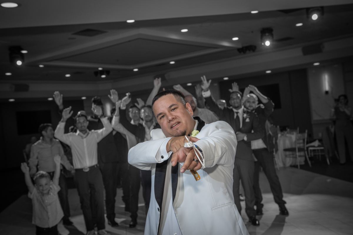 A groom in a white tuxedo points at the camera among celebrating guests, including children and men in suits.