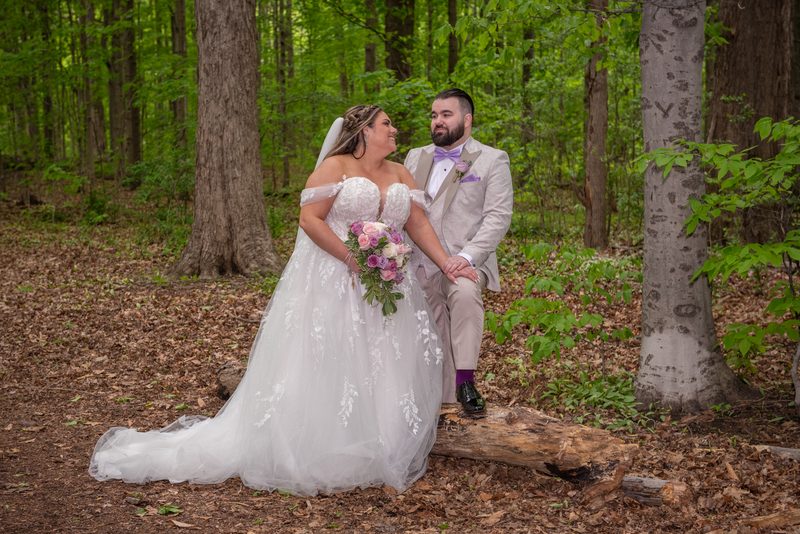 A smiling bride and groom share an intimate moment in a sunlit forest, the bride holding flowers, the groom sitting on a log.