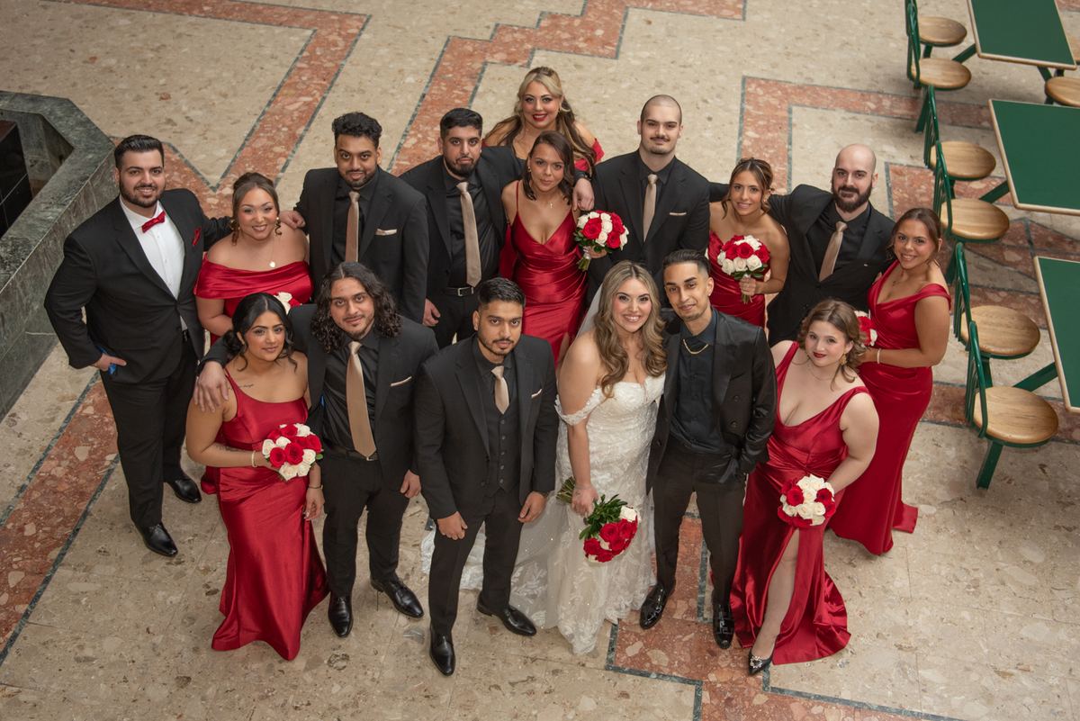 Bride and groom stand with wedding party in an indoor atrium, everyone smiling and holding red-and-white floral bouquets.