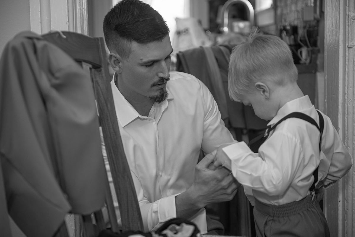 A man adjusts the cuff of a young boy’s shirt in a tailor's shop, with garments hanging nearby, conveying care.