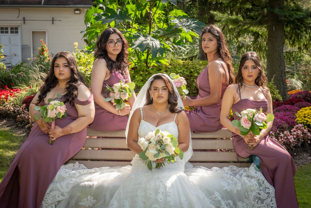 A bride and four bridesmaids pose on a wooden bench surrounded by pink and white roses and green foliage.