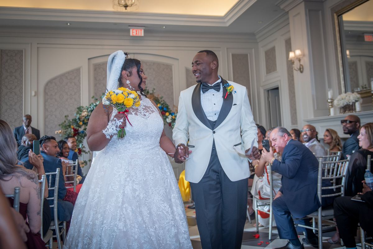 A smiling bride and groom in white walk down the aisle hand-in-hand as guests capture the moment with their phones.