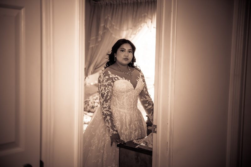 A bride in an elegant lace gown stands thoughtfully by a curtained doorway, illuminated by soft light.