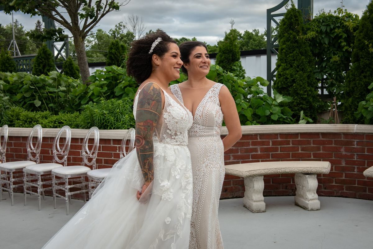 Two brides in white lace gowns smile and gaze joyfully at each other outdoors near a brick patio and greenery.