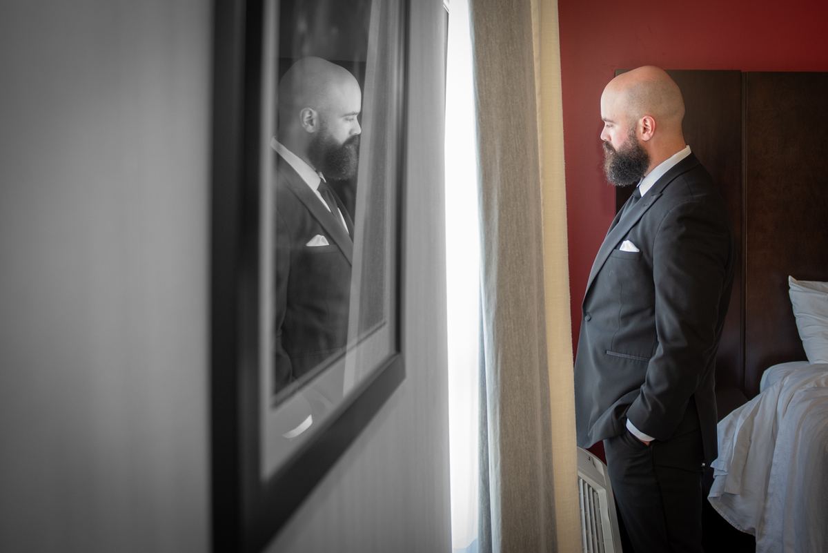 A bearded man in a dark suit stands by a window in a hotel room, his reflection visible in a mirror beside him.