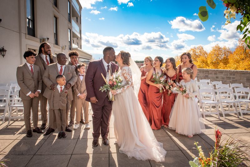 Bride and groom kiss as their wedding party watches joyfully under a blue sky with autumn leaves in the background.