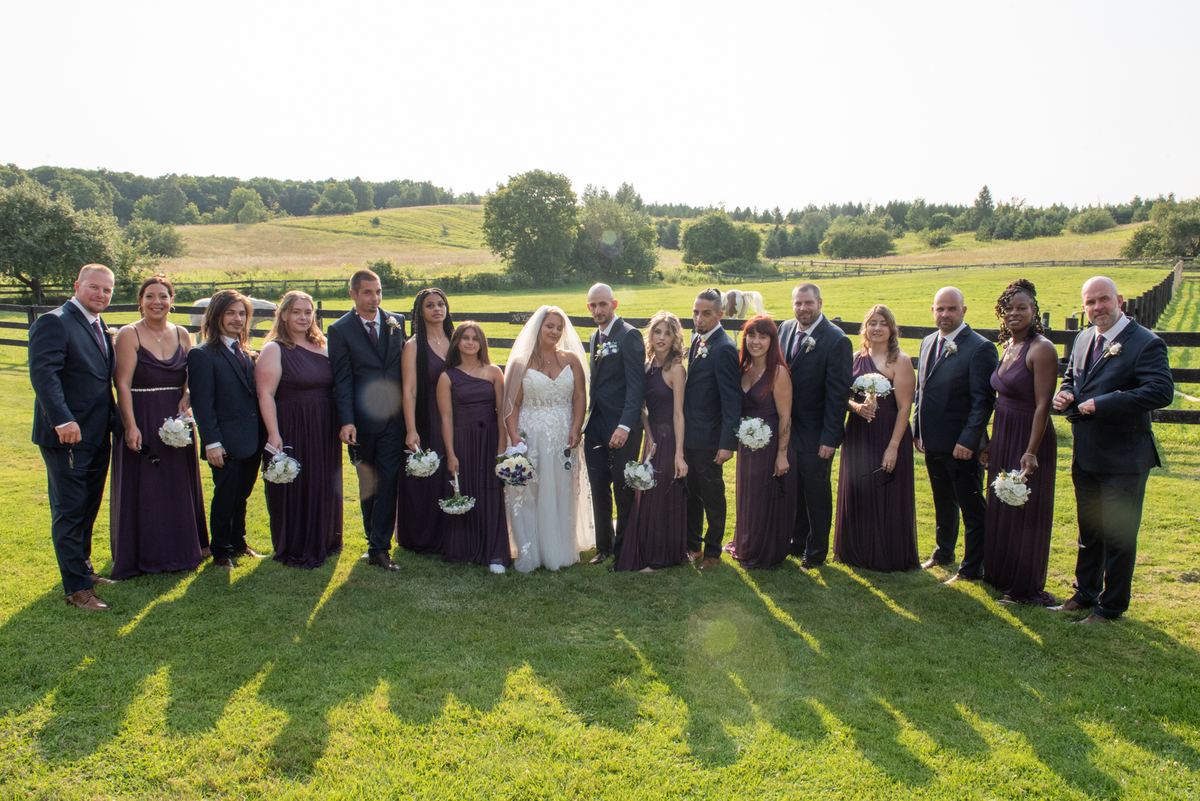 A bride and groom stand with their attendants in coordinated outfits before a wooden fence on a sunny afternoon.