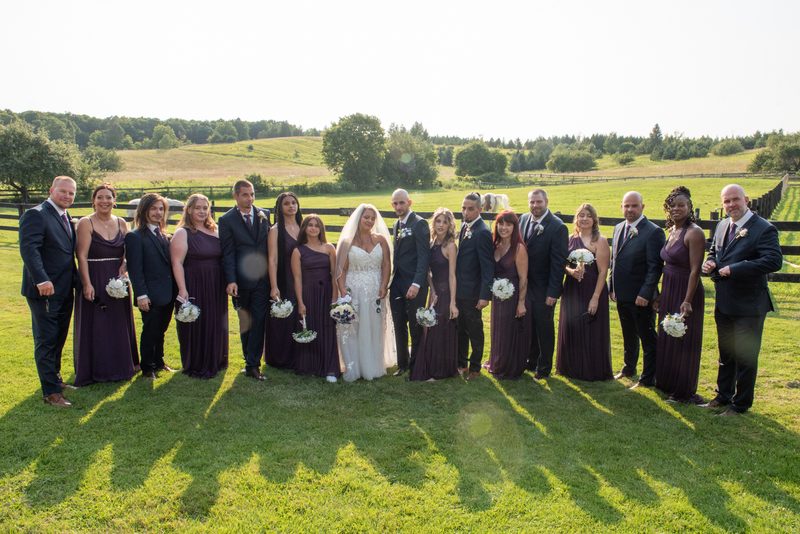 A bride and groom stand with their attendants in coordinated outfits before a wooden fence on a sunny afternoon.
