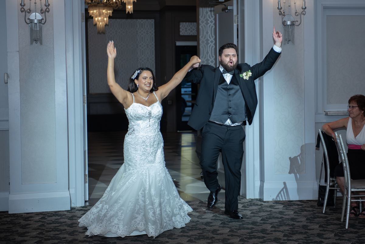 A bride and groom enter a banquet hall, arms raised in joy, as warm light from chandeliers highlights their formal attire.