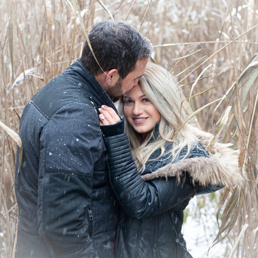 A couple hold each other close among winter reeds as snow falls around them.