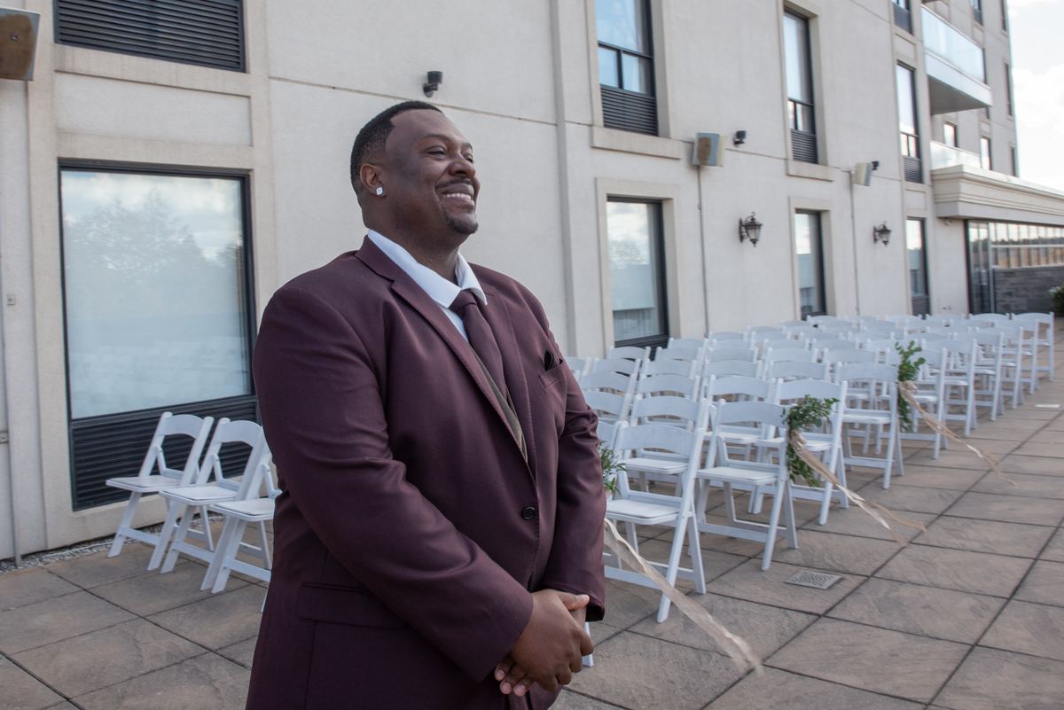 A smiling groom in a burgundy suit stands at the altar, framed by white chairs and greenery, awaiting his bride.