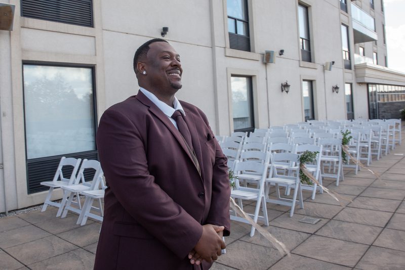 A smiling groom in a burgundy suit stands at the altar, framed by white chairs and greenery, awaiting his bride.