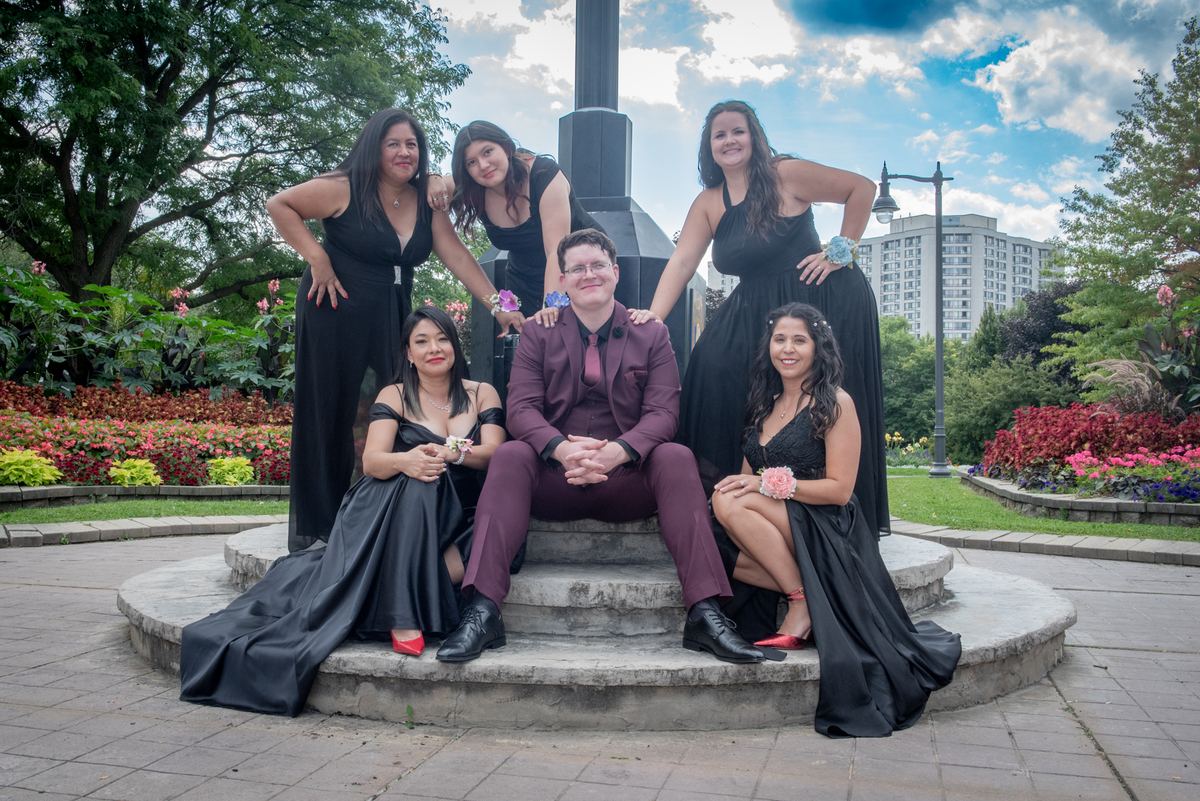Six formally dressed young people pose joyfully on a stone bench in a park with greenery and flowers.
