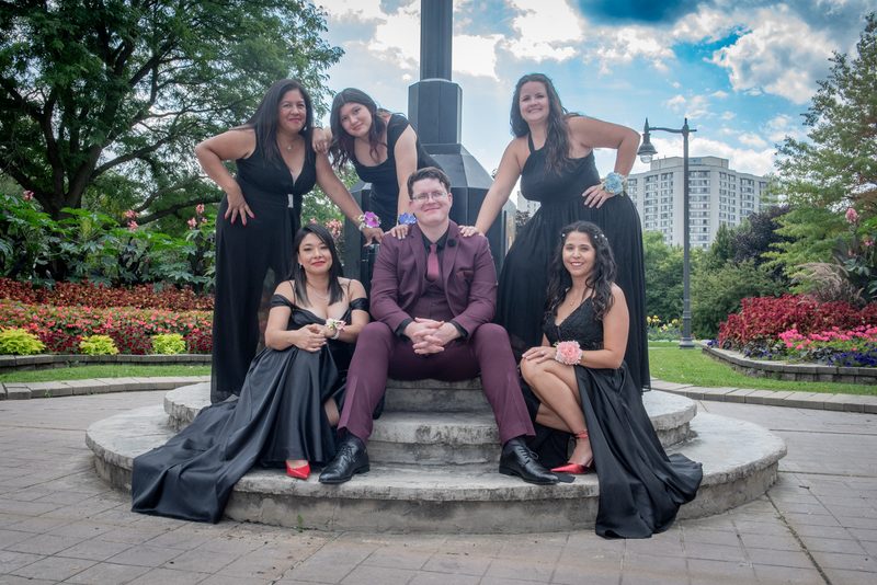 Six formally dressed young people pose joyfully on a stone bench in a park with greenery and flowers.