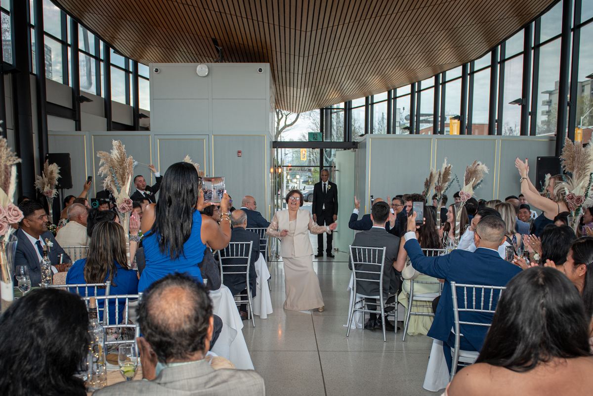 A woman walks down the aisle with outstretched arms as guests cheer and raise phones in a joyful wedding reception.