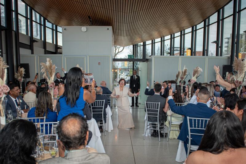 A woman walks down the aisle with outstretched arms as guests cheer and raise phones in a joyful wedding reception.