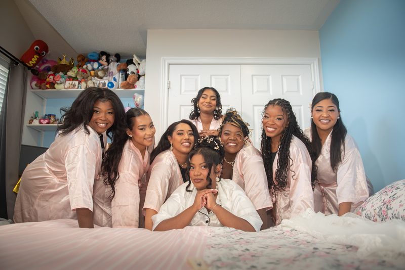 Bride and bridesmaids in matching satin robes smile warmly in a cozy bedroom with stuffed toys on the shelf.