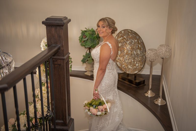 A bride in a lace gown and pink and white roses stands on a curved staircase, smiling at the camera.
