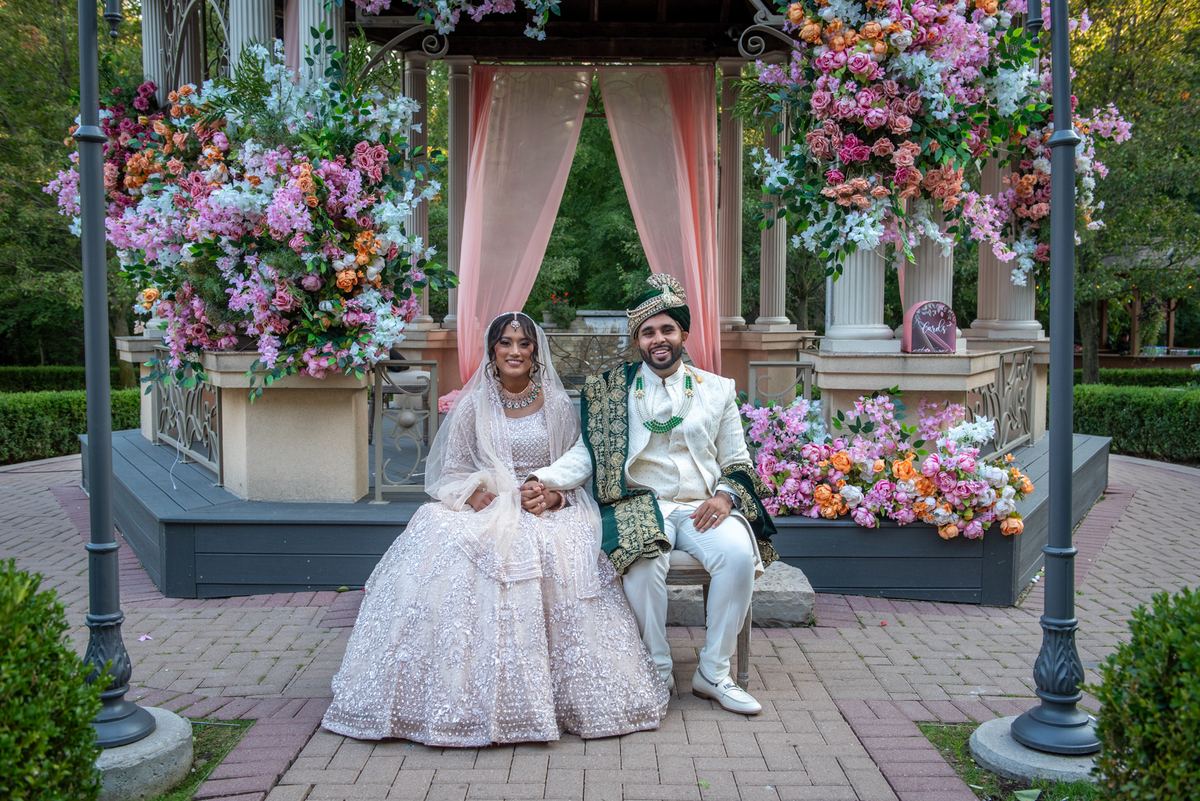 A newlywed couple in traditional South Asian attire sits hand-in-hand in a garden gazebo adorned with colorful flowers.