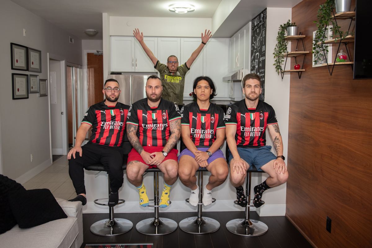 Four people in AC Milan jerseys sit on bar stools in a modern kitchen, one standing behind them with arms raised.