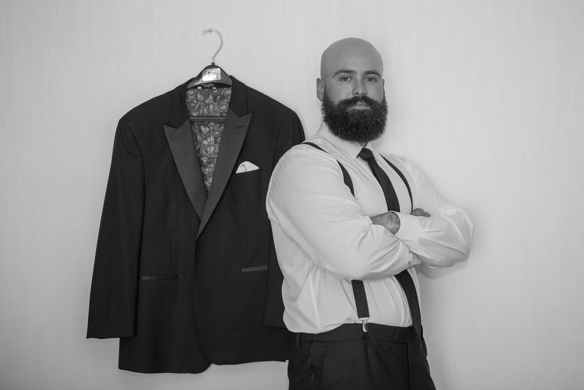 A bearded man in white shirt and suspenders stands beside a tuxedo jacket, suggesting preparation for an event.