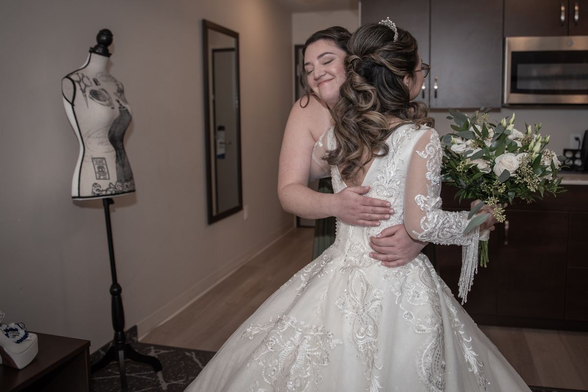 A bride in a white gown embraces a family member, holding flowers near a mannequin with a sketch design.