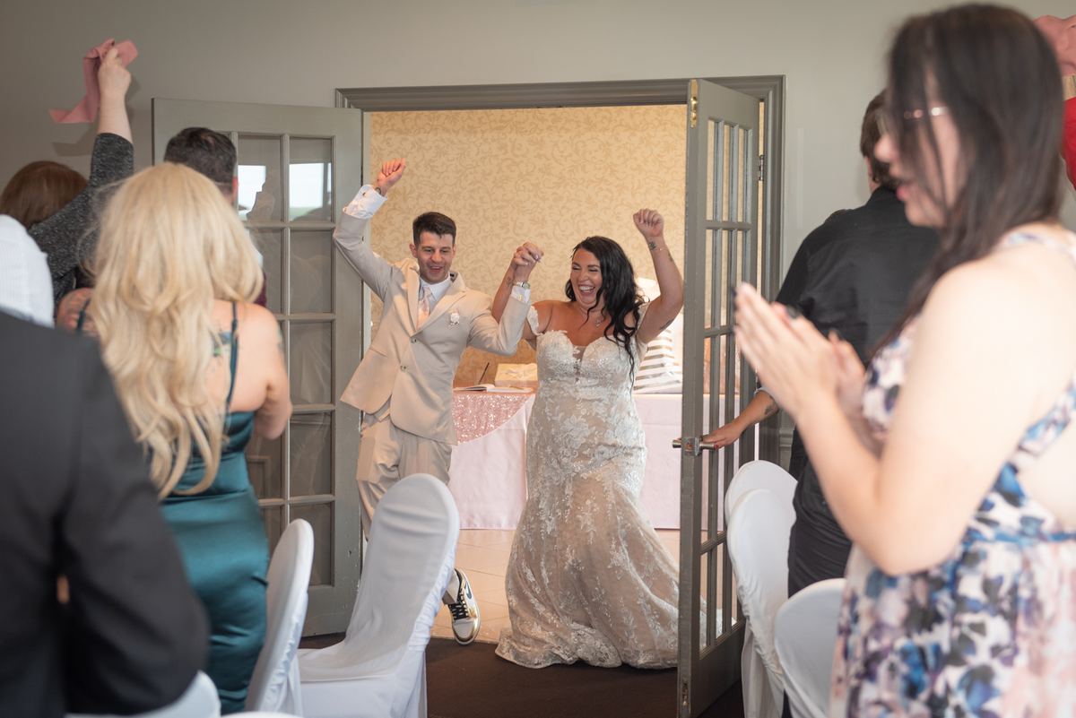 A smiling bride and groom enter a room through double doors, arms raised as guests cheer, celebrating the euphoric moment.