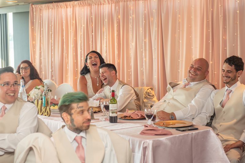 Guests laugh around a banquet table at a wedding reception, illuminated by string lights and drapes.