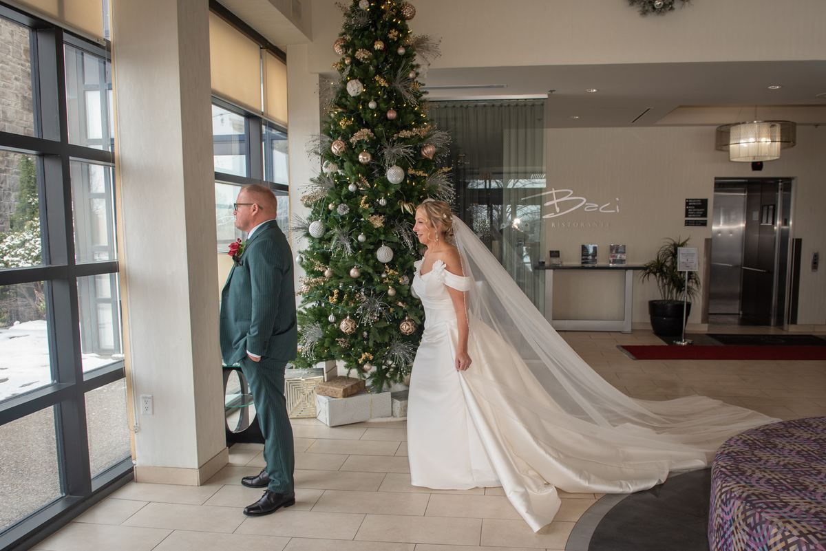 Bride in white gown and veil stands beside groom in teal suit, both looking tenderly at each other near Christmas tree.